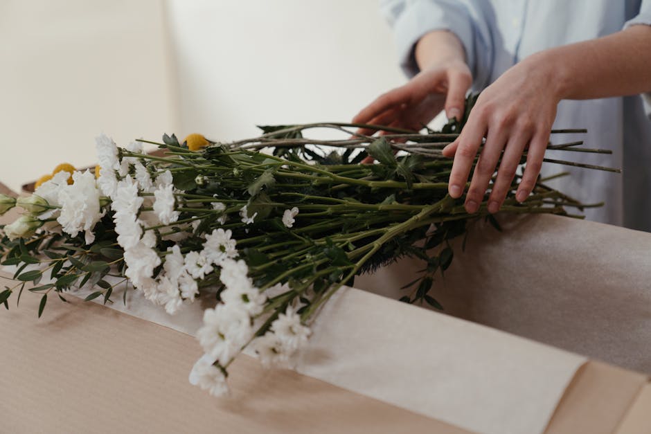 A person’s hands, gently arranging a bouquet of fresh white and yellow flowers, including carnations and daisies, on a light-colored wooden table in a well-lit indoor setting. The bouquet features g