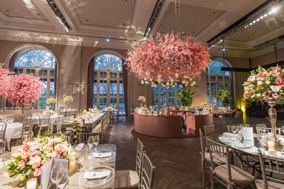 Elegant banquet hall decorated with large floral arrangements featuring pink cherry blossom trees and mixed bouquets of pink roses, peonies, and white blossoms. The flowers are arranged in tall glass 