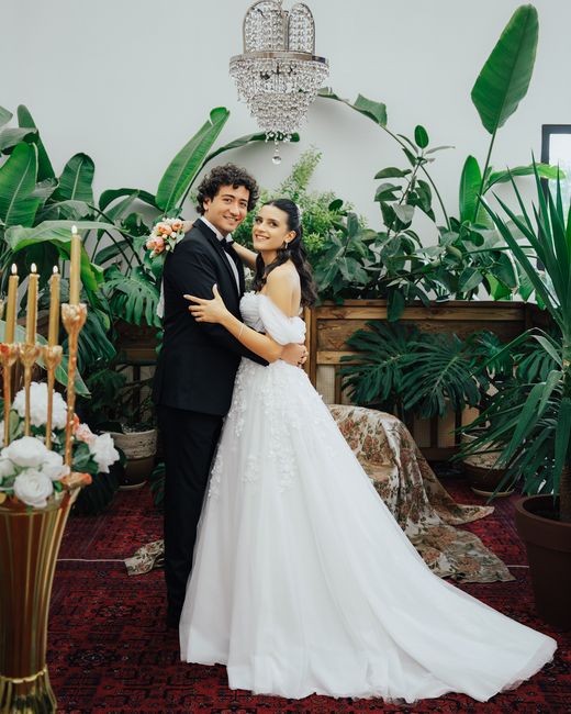A bride and groom in an indoor setting surrounded by lush green tropical plants and large leaves. The bride is wearing a white wedding gown with lace details and a long train, holding a small bouquet 