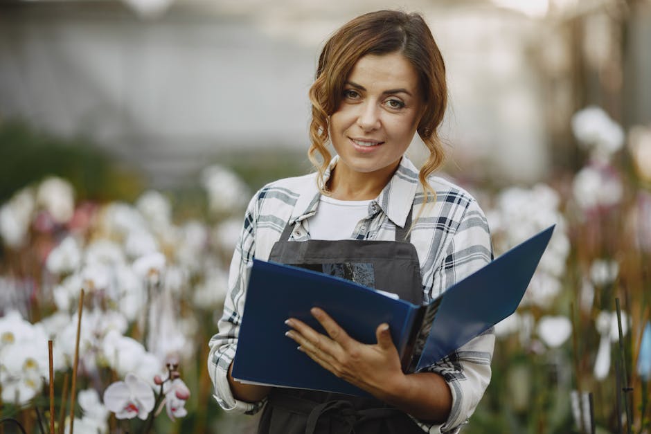 A female florist with wavy, shoulder-length brown hair and light makeup, smiling gently as she holds a blue floral arrangement catalog or price list in her hands. She is dressed in a casual white T-sh
