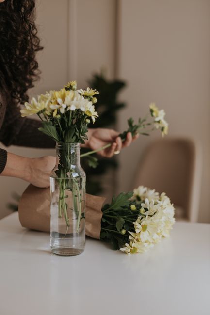 A person with curly hair is arranging a bouquet of fresh white and pale yellow daisies in a transparent glass vase on a white table. The bouquet features vibrant green stems and leaves, with some flow